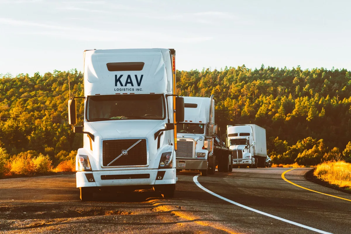 KAV Logistics tractor-trailers lined up on a two-lane road through a forest at golden hour